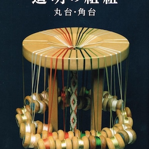 以下が含まれることがあります： 丸い天板と糸巻きの輪が付いた木製の組紐台。台には複数の色の糸があり、上部には「道明の組紐」という日本語のテキストがあります。