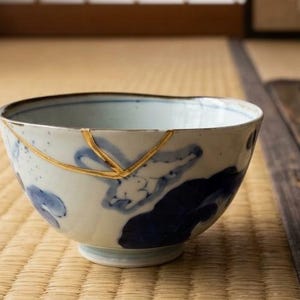 May include: A ceramic bowl with a white and blue floral design, repaired with gold-colored lacquer. The bowl sits on a woven mat, showcasing the Japanese art of Kintsugi.