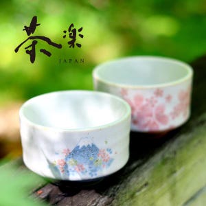May include: Two white ceramic teacups with floral designs. One cup has a mountain scene with blue and pink accents, the other a pink floral pattern. The cups are labelled "茶楽 JAPAN" in black calligraphy.