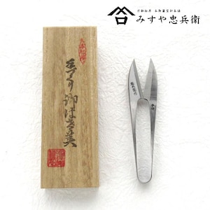 May include: A pair of silver-colored thread snips next to a wooden box. The box has Japanese text and a red stamp. The thread snips have pointed blades and a long handle. The box is light brown with dark brown text.