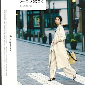 以下が含まれることがあります： クリーム色の服装の女性が横断歩道を歩いています。長いベスト、パンツ、白いシャツを着ています。麦わらトートバッグを持っています。画像には「think pattern」と「ソーイングBOOK」という文字が含まれています。