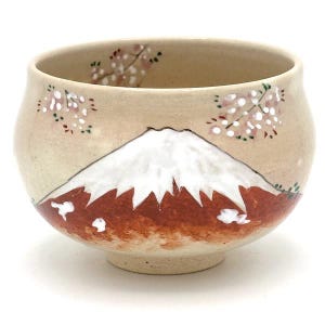 May include: A beige ceramic bowl with a hand-painted design of Mount Fuji and cherry blossoms. The mountain is depicted with a white snow-capped peak and a reddish-brown base. Delicate pink and white blossoms are scattered around the rim.