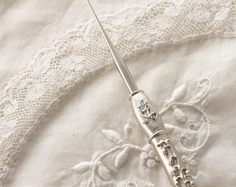 May include: A silver-toned crochet hook featuring an ornate, textured handle with raised decorative scrollwork and a central monogram detail. The metal tool has a smooth, tapered shaft and a curved hook tip, showcasing intricate craftsmanship.