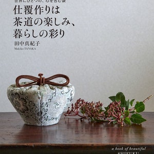 以下が含まれることがあります： 「美しい仕覆: Shifuku」というタイトルの本。日本語で「仕覆作りは茶道の楽しみ、暮らしの彩り」と書かれている。本の表紙には、小さな丸い白と緑の布製のポーチが描かれており、茶色の紐が付けられている。ポーチは、赤い実と緑の葉のついた小枝が置かれた木製のテーブルの上に置かれている。