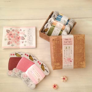 以下が含まれることがあります： 様々な色の刺繍糸の糸巻きが入った蓋付きの木箱。箱は日本の文字と花柄で飾られています。箱が開いていて、糸巻きが見えます。箱の外にも糸巻きが3つあり、2つはピンク、1つは赤です。