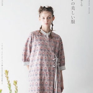 以下が含まれることがあります： 女性が、薄紫色の花柄ラップドレスを着ています。ドレスはマンダリンカラーとタイクロージャーです。画像には、日本語と英語のテキストも含まれており、「Twelve Sewing Book」が含まれています。