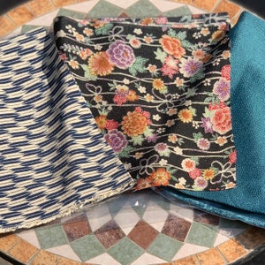 以下が含まれることがあります： 黒い背景に花柄の生地、青と白のヘリンボーン柄の生地、無地のターコイズブルーの生地の3つの生地見本です。