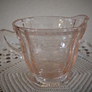 May include: A pink glass creamer with a delicate floral pattern. The creamer has a handle and a footed base.