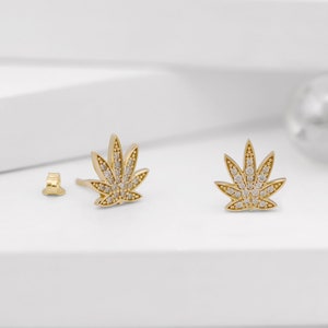 Puede incluir: Un par de pendientes de tono dorado con un diseño de hoja de marihuana. Las hojas están cubiertas de pequeñas piedras de circonita cúbica transparentes.