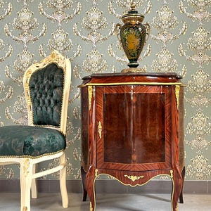 Op de afbeelding: Een antieke houten kast met een donkere, glanzende afwerking en gouden accenten. Een decoratieve groene en gouden vaas staat bovenop de kast. Links staat een witte en gouden stoel met een groen gestoffeerde zitting. De achtergrond is behangen met behang.