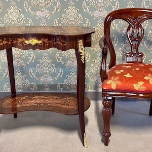 Peut inclure: Une table antique en acajou avec un plateau ovale et une étagère inférieure, avec des incrustations de bois et des accents dorés. Une chaise en bois assortie avec une assise à motifs floraux rouges est à côté de la table. La chaise a un dossier incurvé.
