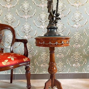 Op de afbeelding: Een kleine, ronde, houten bijzettafel met een donkerbruine afwerking. De tafel heeft een decoratieve rand en een voetstuk. Bovenop staat een bronzen beeld van een vrouw en een cherubijn. Links een houten stoel met een rood patroon.