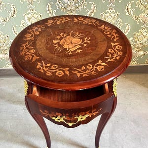 Op de afbeelding: Kleine, ronde houten bijzettafel met een lade. Het tafelblad heeft een ingelegd ontwerp van een muziekinstrument en bloemmotieven. De tafel heeft gebogen poten met gouden accenten en de lade is open.