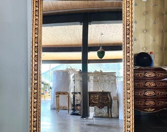 May include: A large, rectangular gold-framed mirror with ornate floral detailing at the top and along the edges. The mirror reflects a room with furniture and a patterned wallpaper background. The frame has a warm, antique appearance.