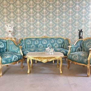 Può includere: Un lussuoso set da soggiorno con un divano e due poltrone con motivo floreale turchese e finiture dorate. Un tavolino abbinato si trova di fronte al divano. La stanza ha una carta da parati a motivi e un vaso decorativo.