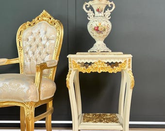 May include: An ornate, cream-colored side table with gold accents and a matching chair. A decorative vase with floral designs sits atop the table. The chair and table feature intricate carvings and a classic, elegant design.