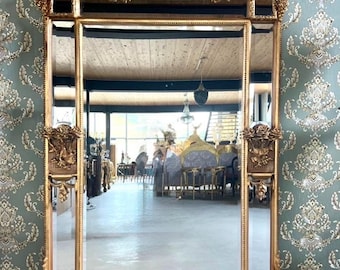 May include: A large, ornate gold-framed mirror with intricate floral and scroll detailing. The rectangular mirror has a decorative top and base, with smaller rectangular mirrors on either side. The mirror is a statement piece, reflecting a room with antique furniture.