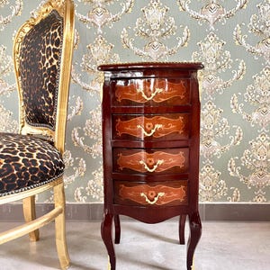 Könnte beinhalten: Ein kleiner, dunkelbrauner Holzschrank mit vier Schubladen, mit dekorativen floralen Intarsien und goldfarbener Hardware. Der Schrank hat geschwungene Beine mit goldenen Akzenten. Links steht ein goldgerahmter Stuhl mit Leopardenmuster.