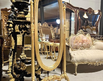 May include: A gold-colored, oval-shaped floor mirror with ornate detailing. The mirror is supported by a decorative stand with curved legs. The frame and stand feature intricate carvings, reflecting a classic, antique style. Other antique furniture is visible in the background.