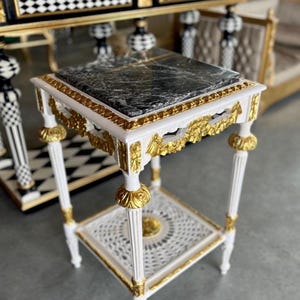 May include: An ornate side table with a white frame, gold accents, and a black marble top. The table has a decorative lower shelf and measures approximately 50cm in height.