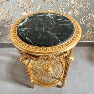 Op de afbeelding: Een kleine, sierlijke bijzettafel met een ronde, donkergroene marmeren bovenkant. De tafel heeft een goudkleurig, gesneden houten frame met een onderste plank en decoratieve details. De tafel is ongeveer 61 cm hoog en 51 cm in diameter.