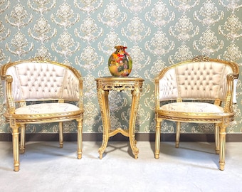 May include: Two gold-framed armchairs with tufted cream-colored backs and seats flank a small, ornate gold table. A colorful vase sits atop the table. The chairs and table have detailed carvings, and the background features a patterned wallpaper.