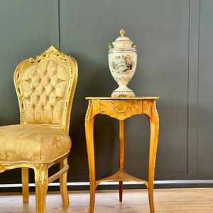 May include: A gold-framed chair with a tufted, patterned seat sits next to a small, ornate wooden table. A decorative vase with a lid, featuring painted scenes, rests on the table. The chair and table have a vintage aesthetic.