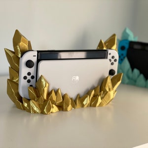 May include: A white Nintendo Switch console is displayed in a gold crystal-shaped stand. The stand is made of 3D printed plastic and features a detailed design of sharp, pointed crystals.