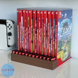 Puede incluir: Un soporte de cartuchos de videojuegos de color marrón en forma de ladrillo con 16 ranuras. El soporte está lleno de varios cartuchos de juegos de Nintendo Switch, incluyendo The Legend of Zelda: Link's Awakening, Breath of the Wild, Tears of the Kingdom, Skyward Sword HD, Mario Party Superstars, Super Mario Party, Mario Kart 8 Deluxe, Mario Golf: Super Rush, Mario Tennis Aces, Super Mario Odyssey, Super Mario 3D World Bowser's Fury y Super Mario 3D All-Stars.