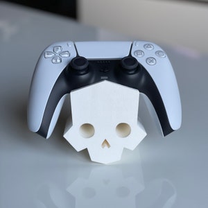 May include: A white PlayStation 5 DualSense controller sits on a white skull-shaped stand. The controller has black buttons and a black faceplate.