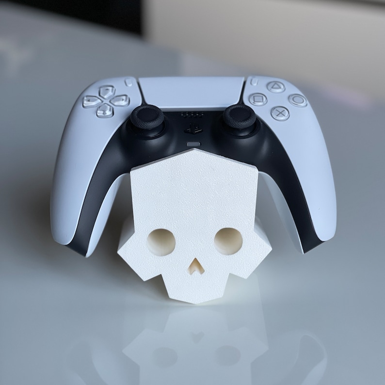May include: A white PlayStation 5 DualSense controller sits on a white skull-shaped stand. The controller has black buttons and a black faceplate.