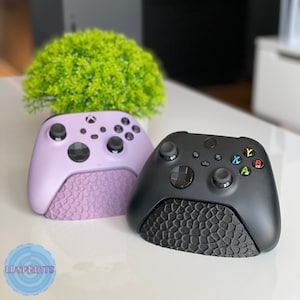 Puede incluir: Dos mandos de Xbox, uno morado y otro negro, se muestran sobre una superficie blanca. Ambos mandos descansan sobre una base texturizada.