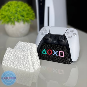 Könnte beinhalten: Ein weißer PlayStation 5 Controller-Ständer mit schwarzer Basis und den PlayStation-Knopfsymbolen in verschiedenen Farben. Der Ständer besteht aus einem strukturierten Material und ist so konzipiert, dass er einen PlayStation 5 Controller hält.