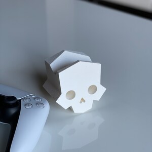 May include: A white, 3D printed skull-shaped phone stand. The stand has a geometric design and is made of a smooth, white plastic.