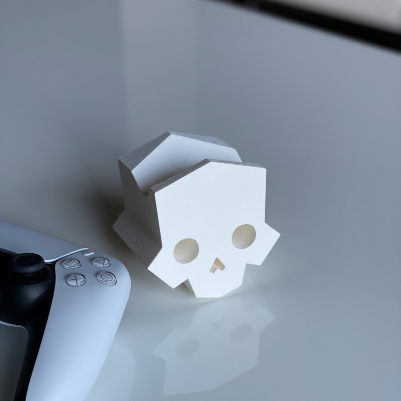May include: A white, 3D printed skull-shaped phone stand. The stand has a geometric design and is made of a smooth, white plastic.