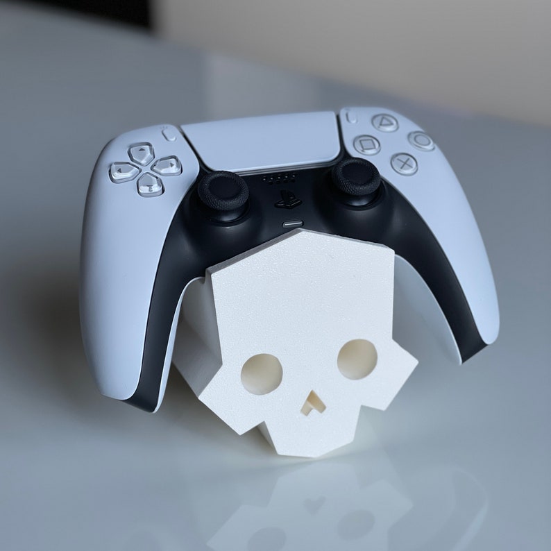 May include: A white PlayStation 5 DualSense controller with black buttons and a skull-shaped stand. The controller is resting on the stand, which is made of white plastic.