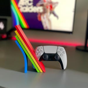 May include: A white and black games controller sits next to a multi-coloured stand. The stand has layers of blue, green, yellow, and red. A television screen with the text "ARC Raiders" is in the background.