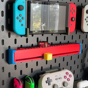 Puede incluir: Un panel perforado negro con varios accesorios de juego. Una consola Nintendo Switch con mandos azules y rojos está montada. Debajo, un soporte rojo para cartuchos de juego con un bloque de signo de interrogación amarillo. También se muestran otros accesorios.