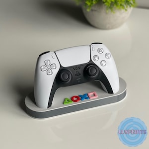 Puede incluir: Un mando de PlayStation 5 blanco con detalles negros se muestra en un soporte gris. El soporte tiene una base blanca con símbolos de PlayStation de colores.