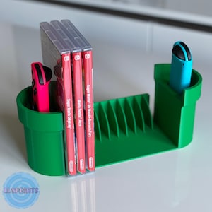 Może przedstawiać: Zielony stojak na gry i kontrolery Nintendo Switch, wydrukowany w 3D w kształcie rury z Super Mario Bros. Stojak pomieści 12 kartridży z grami i ma dwa gniazda na kontrolery. Stojak jest pokazany z trzema pudełkami na gry i dwoma kontrolerami.