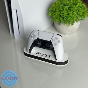 Puede incluir: Un mando de PlayStation 5 blanco se encuentra en una base de carga blanca y negra con el logotipo de PlayStation 5.