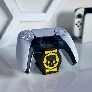 Può includere: Un controller bianco PlayStation 5 DualSense è posizionato su un supporto nero con un logo di teschio giallo. Il controller è in posizione verticale con i pulsanti rivolti in avanti.