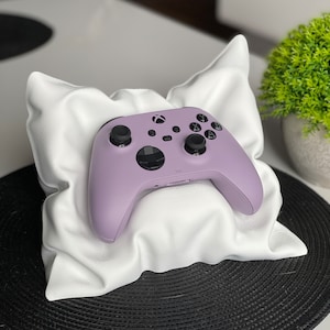 Pode incluir: Um controlador Xbox roxo-lavanda com botões pretos está descansando em um travesseiro branco. O travesseiro está em uma superfície preta.