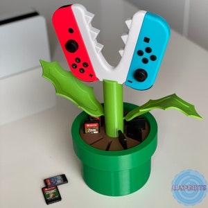 May include: A 3D printed plant pot shaped like a Piranha Plant from the Super Mario Bros. video game. The pot is green and has two white and red Nintendo Switch controllers for the plant's mouth. The pot has a brown base and is filled with slots for game cartridges.