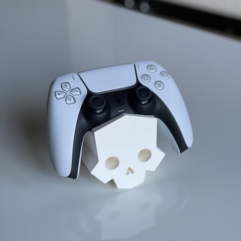 May include: A white PlayStation 5 controller is resting on a white skull-shaped stand. The controller has black buttons and a black PlayStation logo.