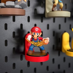 Peut inclure: Figurine colorée de Mario, le personnage emblématique de jeu vidéo, dans une pose dynamique avec un effet de feu. La figurine est montée sur une base rouge et placée sur un fond de panneau perforé noir. D'autres figurines de personnages de jeux sont également visibles.