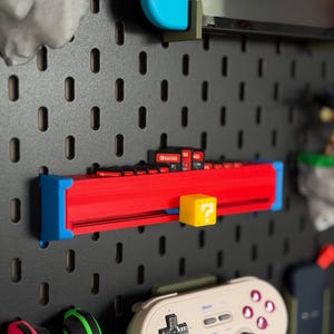 Puede incluir: Un soporte rojo y azul para cartuchos de juego con un bloque amarillo de signo de interrogación, montado en una panel perforado negro. Se ve un mando de juego azul, junto con otros accesorios de juego. El soporte está diseñado para organizar cartuchos de juego.