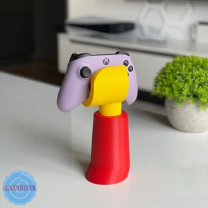 Pode incluir: Um controlador de videogame roxo e preto está montado em um suporte amarelo e vermelho. O suporte tem uma base redonda e uma forma cilíndrica.