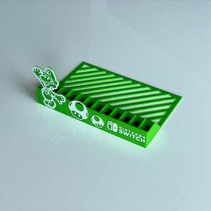 Può includere: Portacarte per giochi Nintendo Switch in plastica verde con un design a strisce bianche. Il portacarte presenta una silhouette bianca di Mario e un fungo. Il testo "NINTENDO SWITCH" è stampato sul lato.