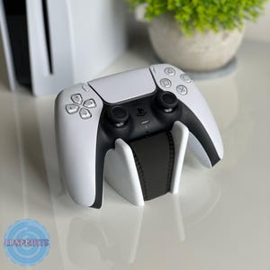 Peut inclure: Une manette de jeu PlayStation 5 DualSense blanche est montée sur un support de charge blanc. La manette a des boutons noirs et un logo PlayStation noir.